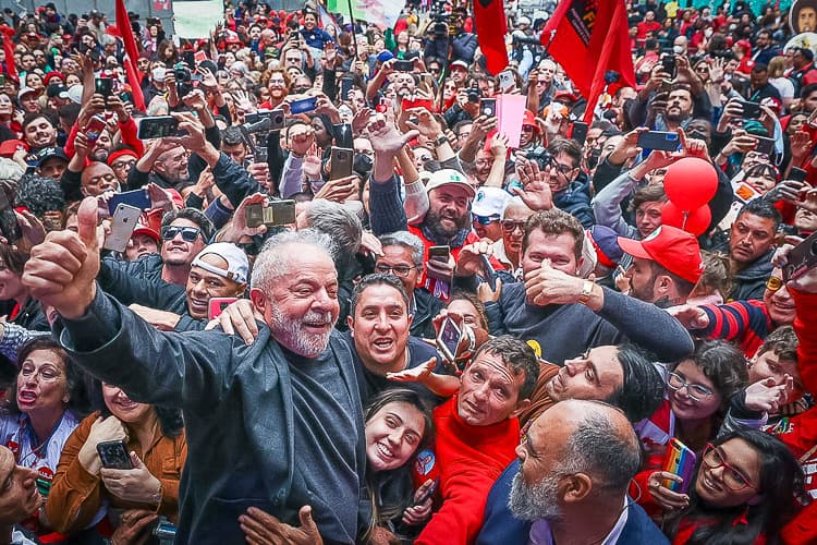 Lula sorri e faz sinal de positivo, cercado por apoiadores com celulares, bandeiras vermelhas e balões em ato de campanha